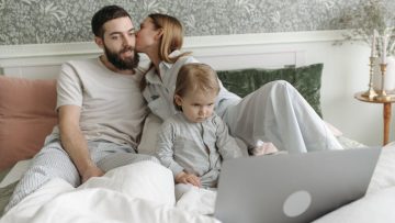 Parents with their child enjoying a morning routine together in a cozy bedroom setting, watching a laptop.