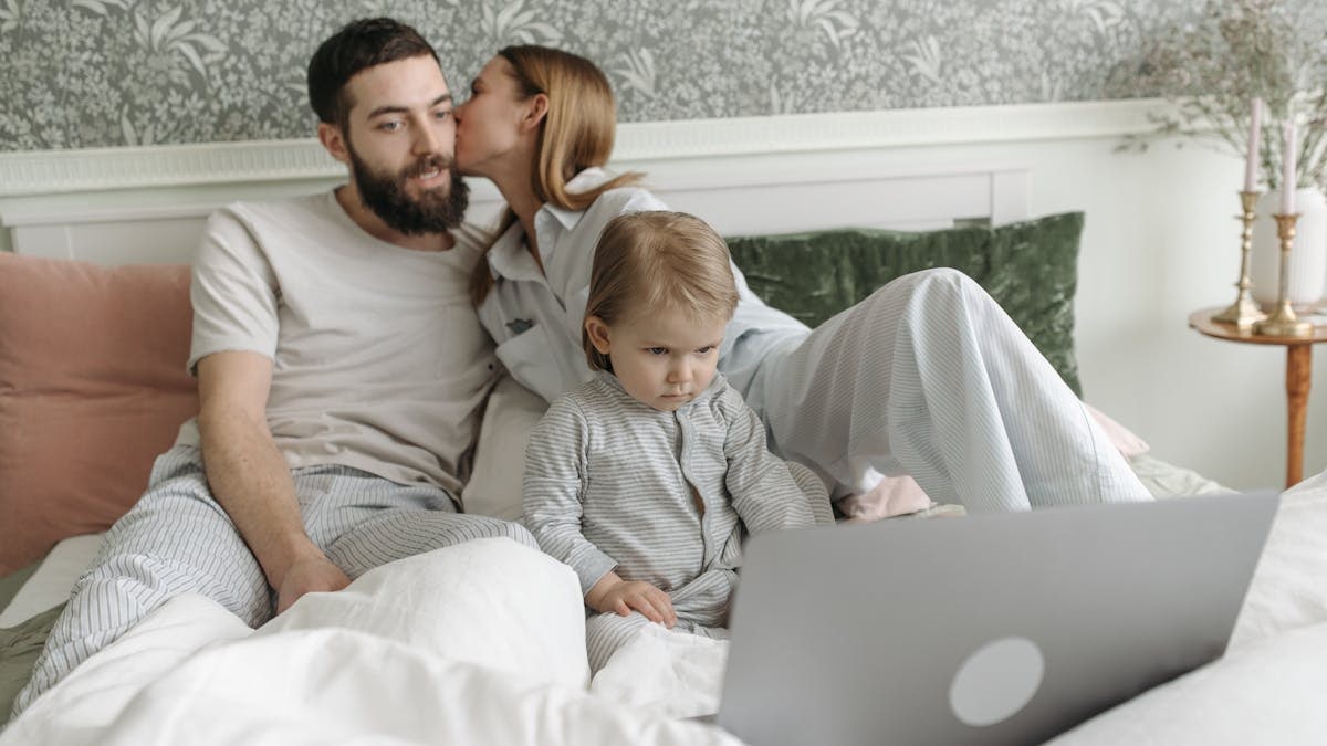 Parents with their child enjoying a morning routine together in a cozy bedroom setting, watching a laptop.